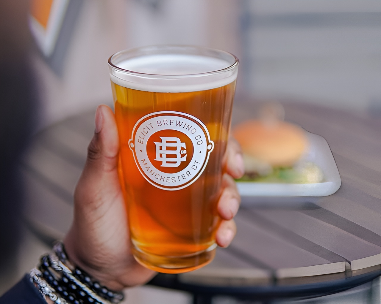 A hand holds a glass of Otravesa Mexican Lager by Elicit Brewing with Cali Love Burger blurred in the background.