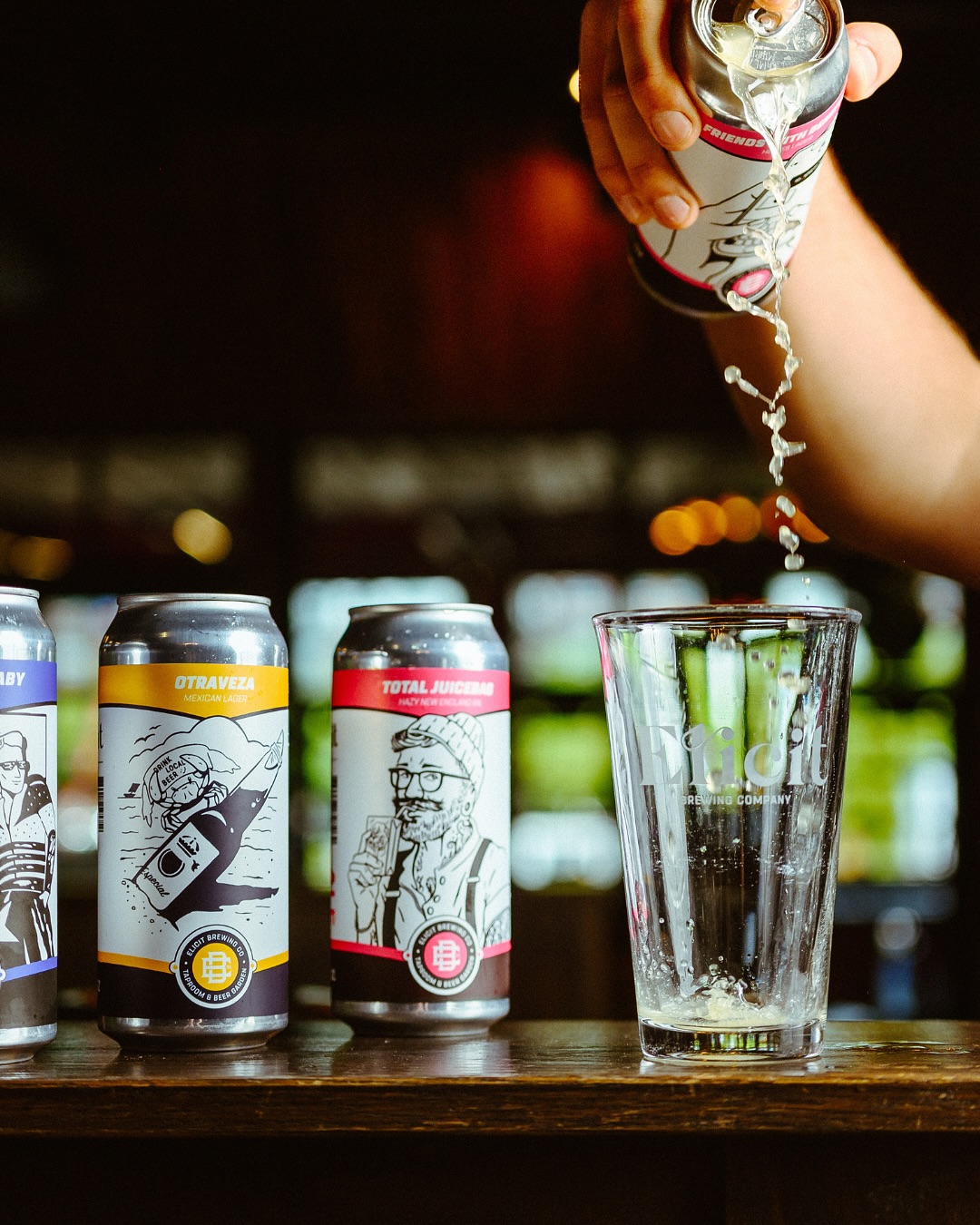 Beer being poured from an Elicit Brewing beer can into a branded pint glass, with several beer cans lined up.