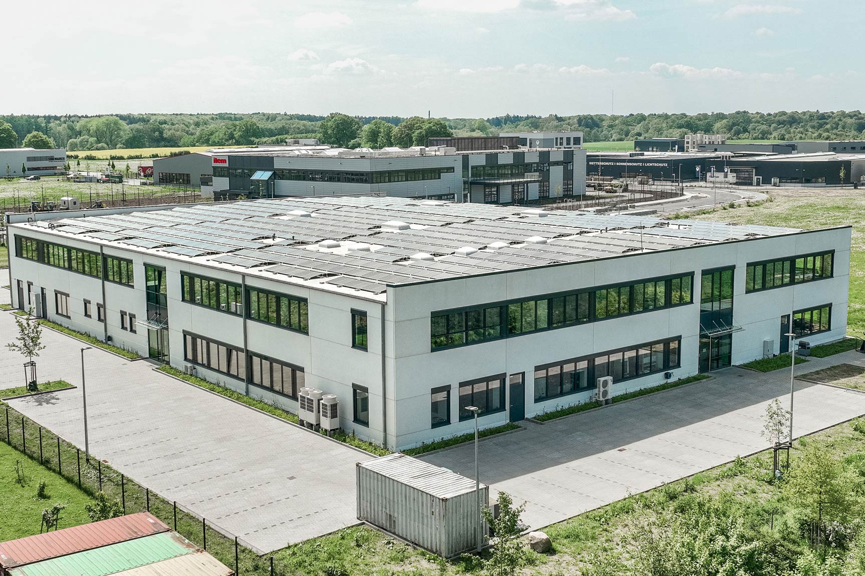 Helles, dreistöckiges Bürogebäude mit Produktionshalle und parkenden Autos davor.