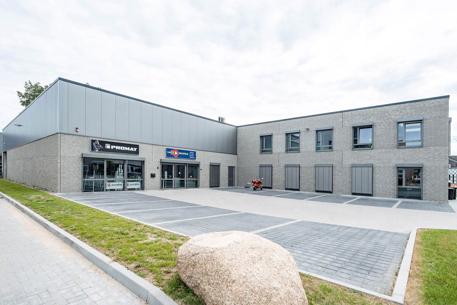 Büro- und Verwaltungsgebäude mit angrenzender Verkaufshalle, hervorgehoben durch einen großen Stein im Vordergrund.