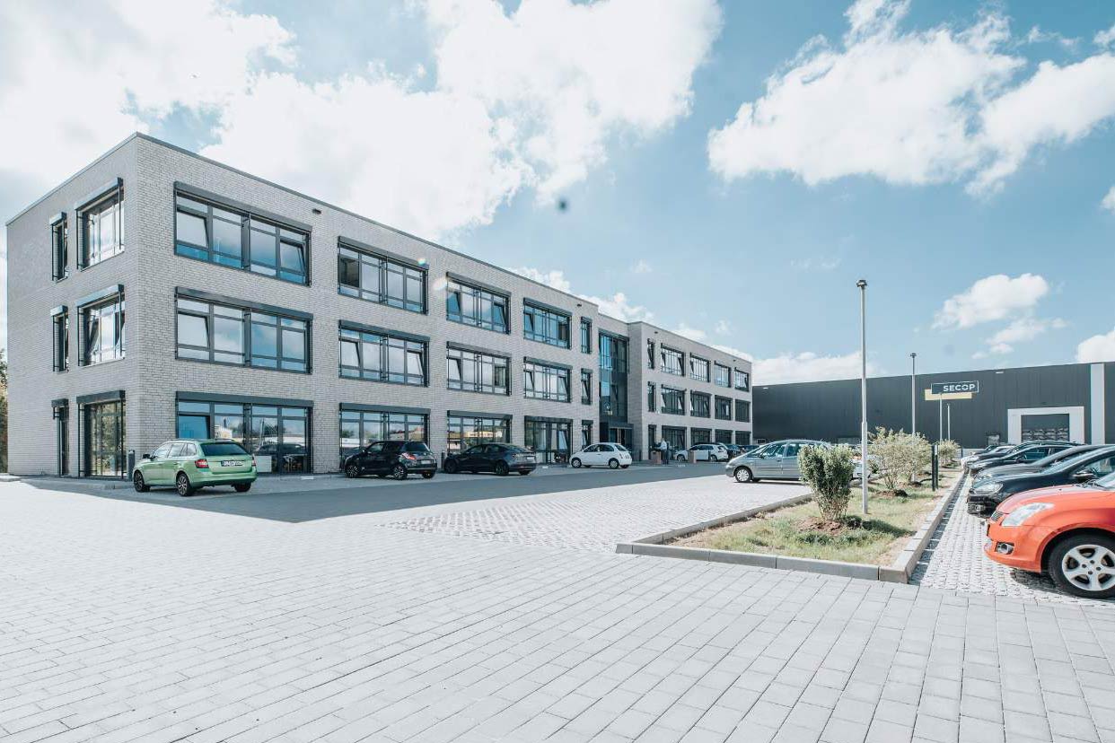 Helles, dreistöckiges Bürogebäude mit Produktionshalle und parkenden Autos davor.