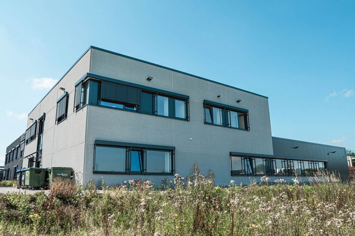 Helles, dreistöckiges Bürogebäude mit Produktionshalle und parkenden Autos davor.