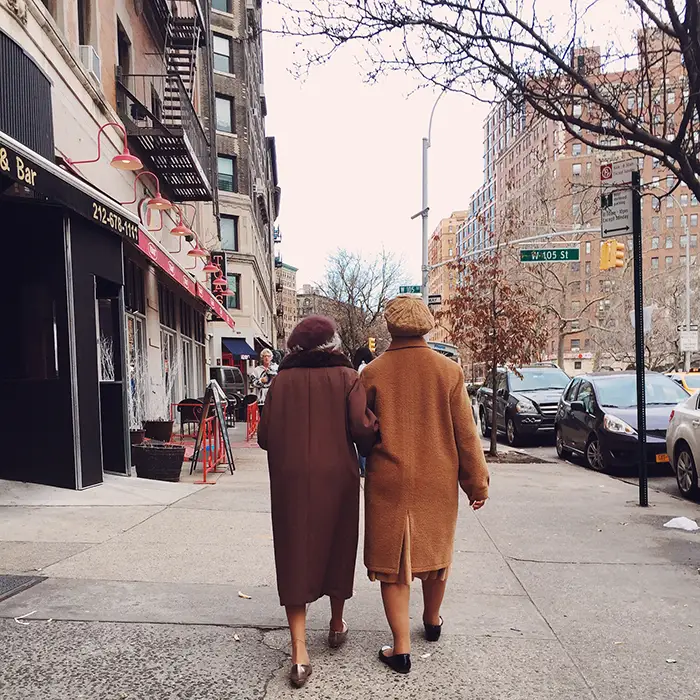 Two older women in peacoats with arms linked walking away from the camera on city street