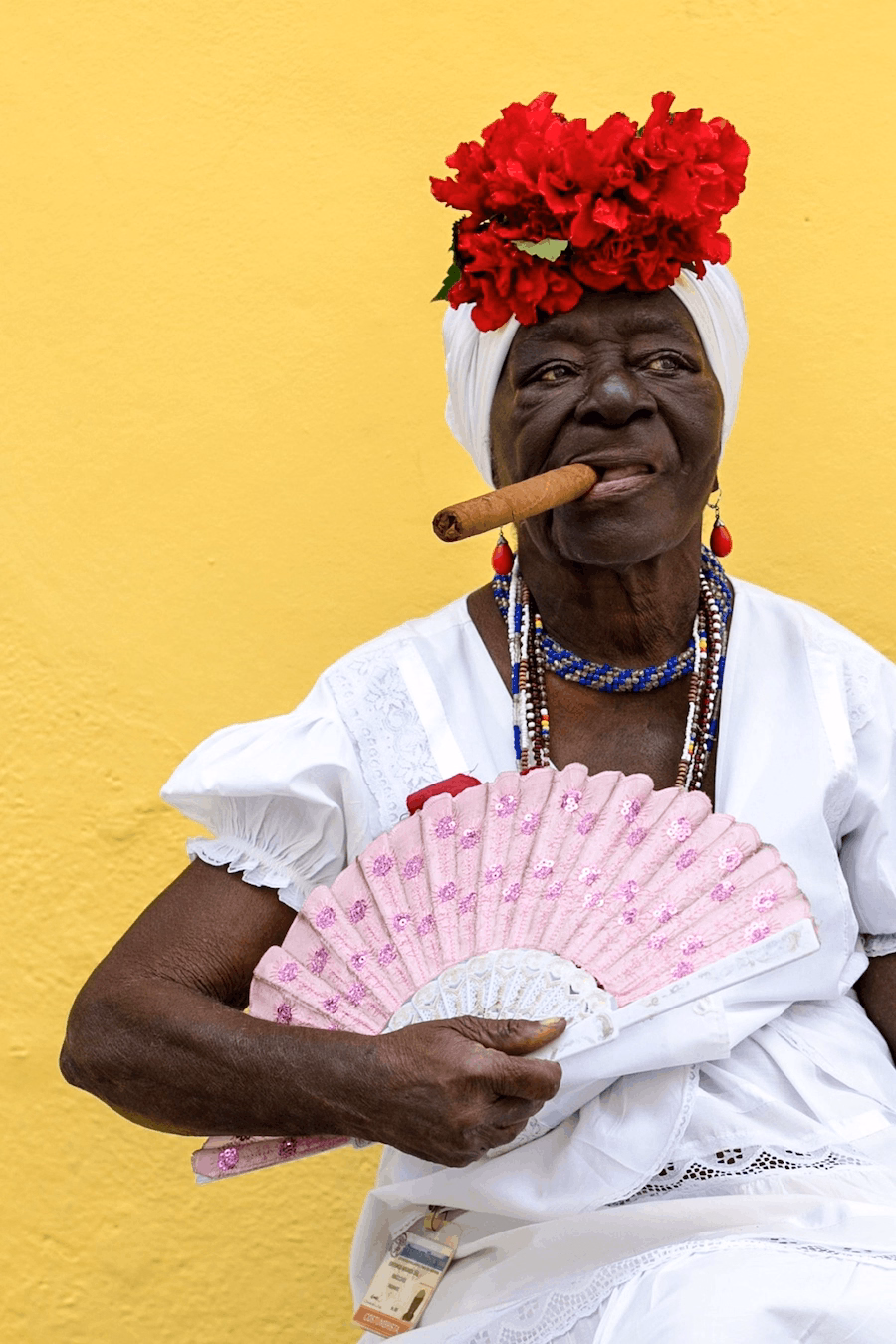 Vibrant portrait photo of a person with red flowers on head holding a pink fan and smoking a cigar in front of a bright yellow wall taken by Liza Gershman