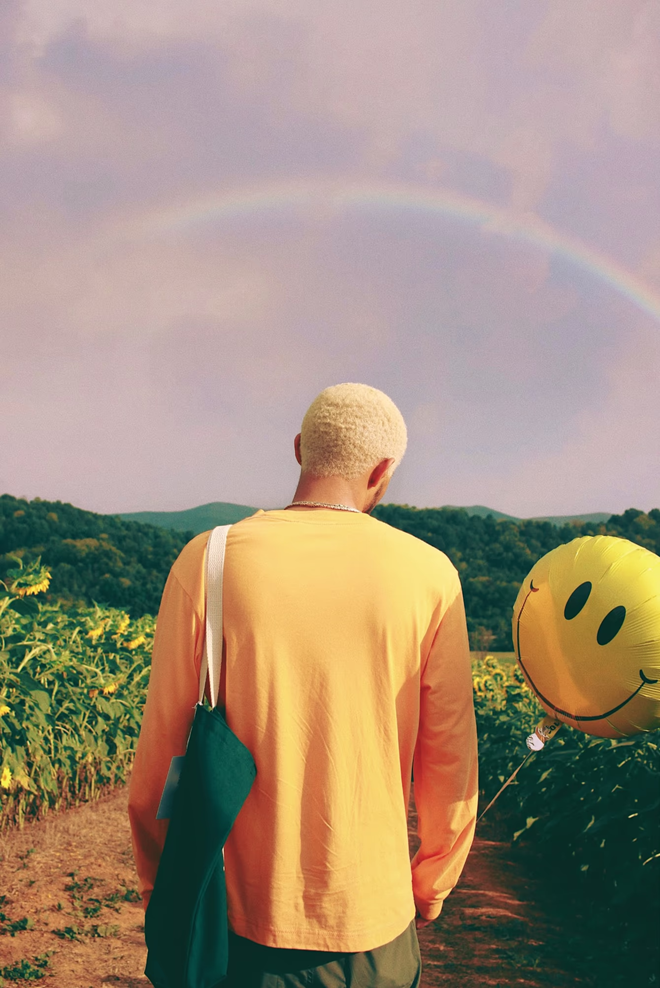 Tess Burris photo of the back of person wearing a yellow long sleeve holding a yellow smiley face ballon in a sunflower field with a rainbow in the sky