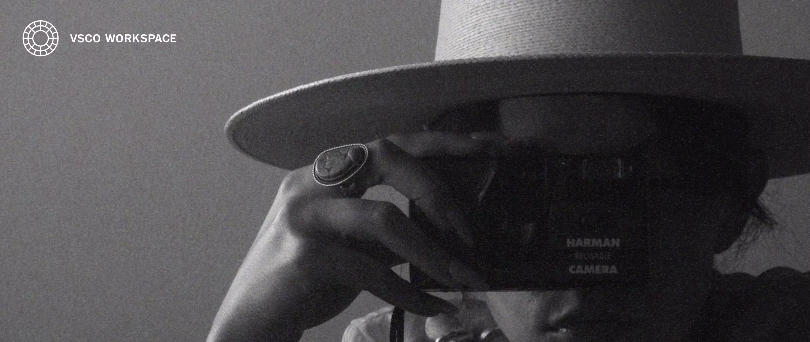 Retrato em preto e branco de uma pessoa de chapéu de aba larga segurando uma câmera de filme diante do rosto, com o logotipo do VSCO Workspace