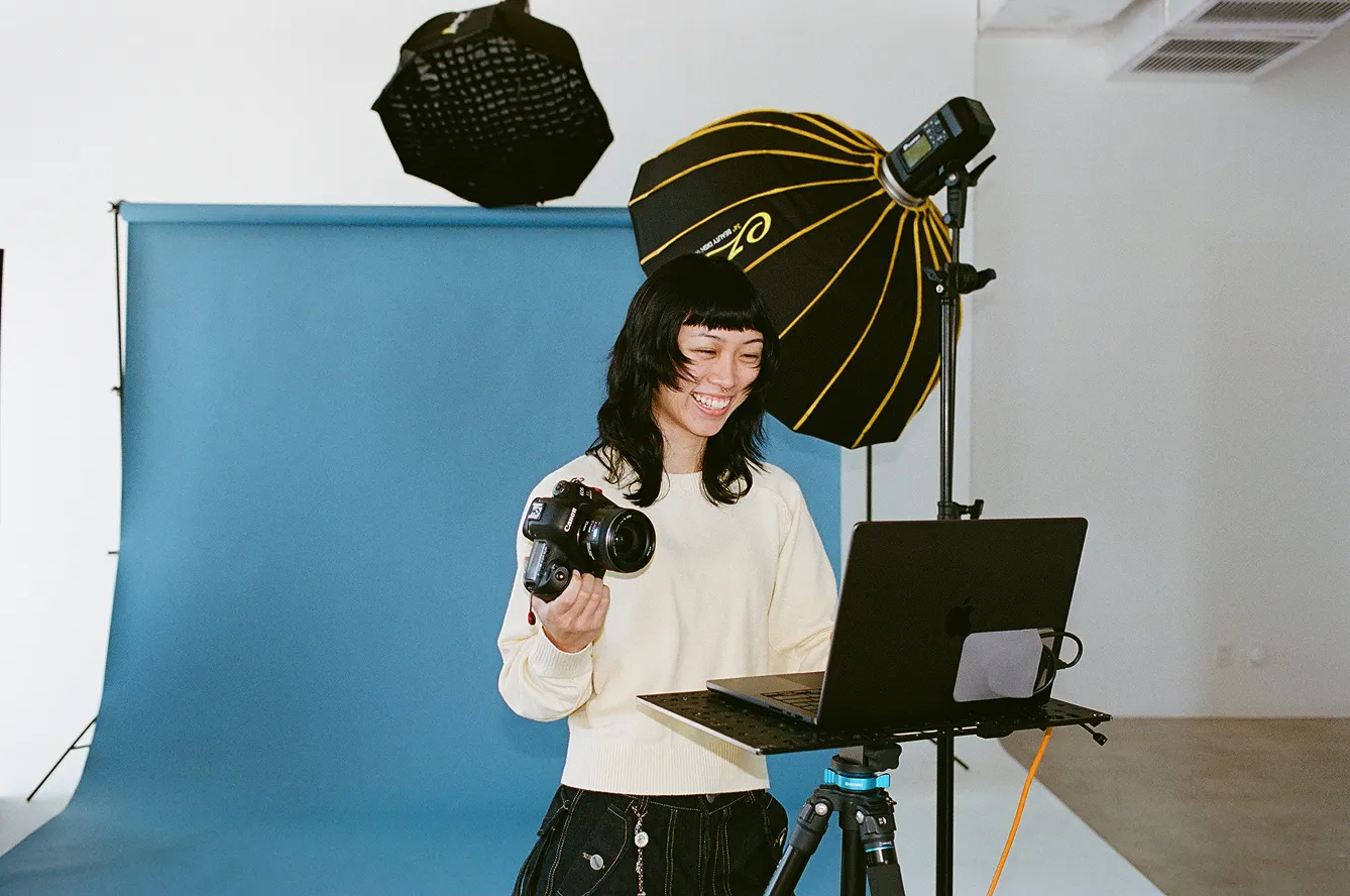 Photographer on set of a brand photoshoot holding a camera, smiling, and looking at a laptop