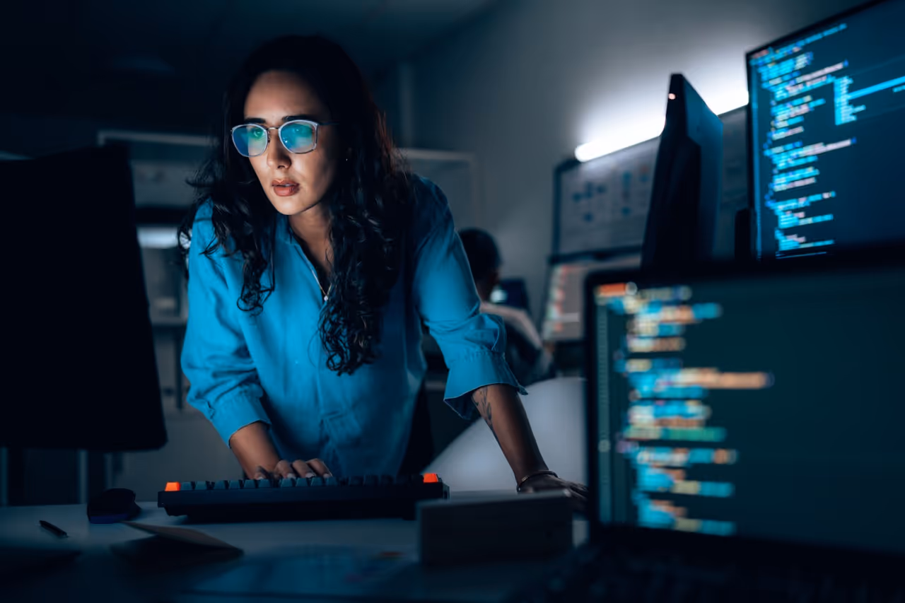 Woman with glasses working late on multiple computer screens displaying code in a dark office.