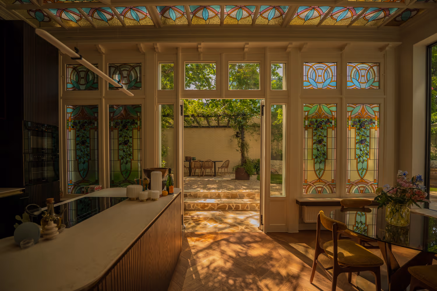 Zonovergoten keuken met glas-in-loodramen en -plafond, die uitkomt op een tuinpatio met stoelen. Warme, rustige sfeer en elegant design.