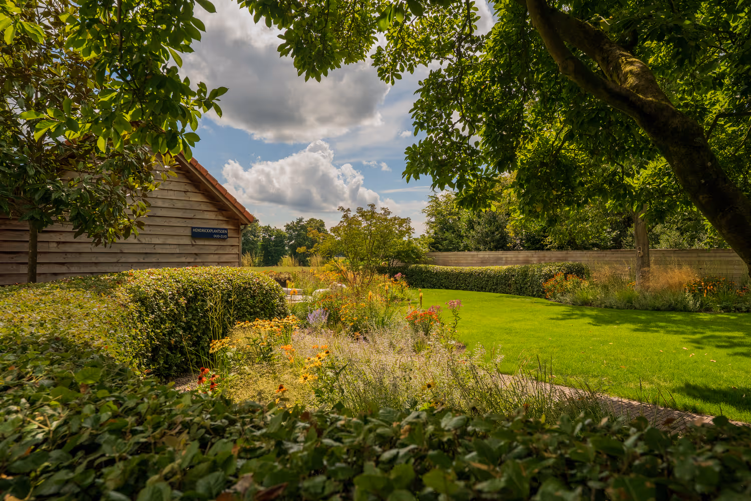 Een weelderige tuin met kleurrijke bloemen en verzorgde struiken onder een gedeeltelijk bewolkte hemel. Links staat een houten gebouw, wat een rustig, zonnig tafereel creëert.