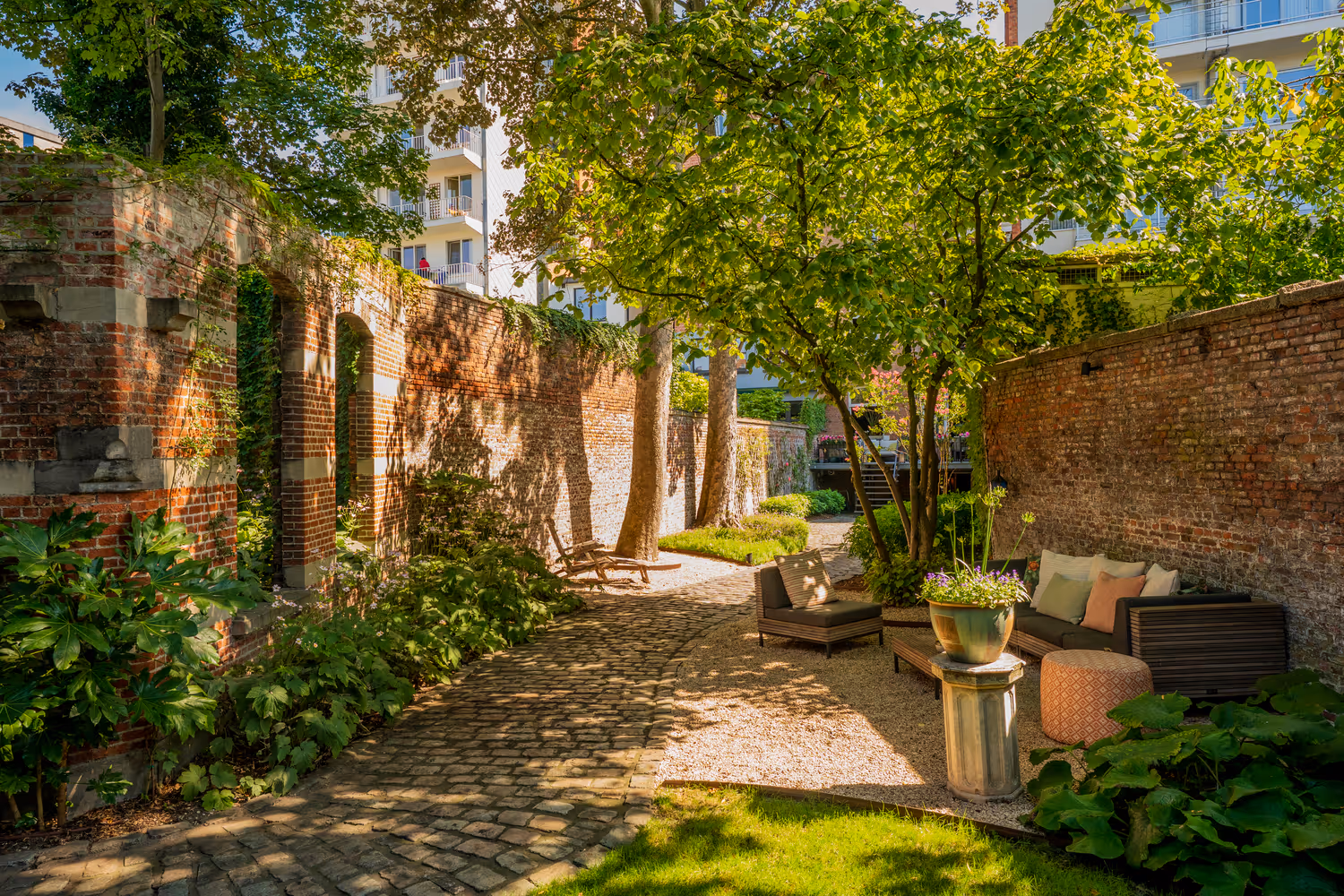 Rustige tuin met geplaveid pad, weelderige bomen en bakstenen muren. Gezellige bank en stoelen creëren een serene zithoek. Zonlicht filtert door de bladeren.