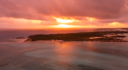 Aerial view of a coastal landscape at sunset with orange and pink clouds reflecting on calm water near dark landmasses.