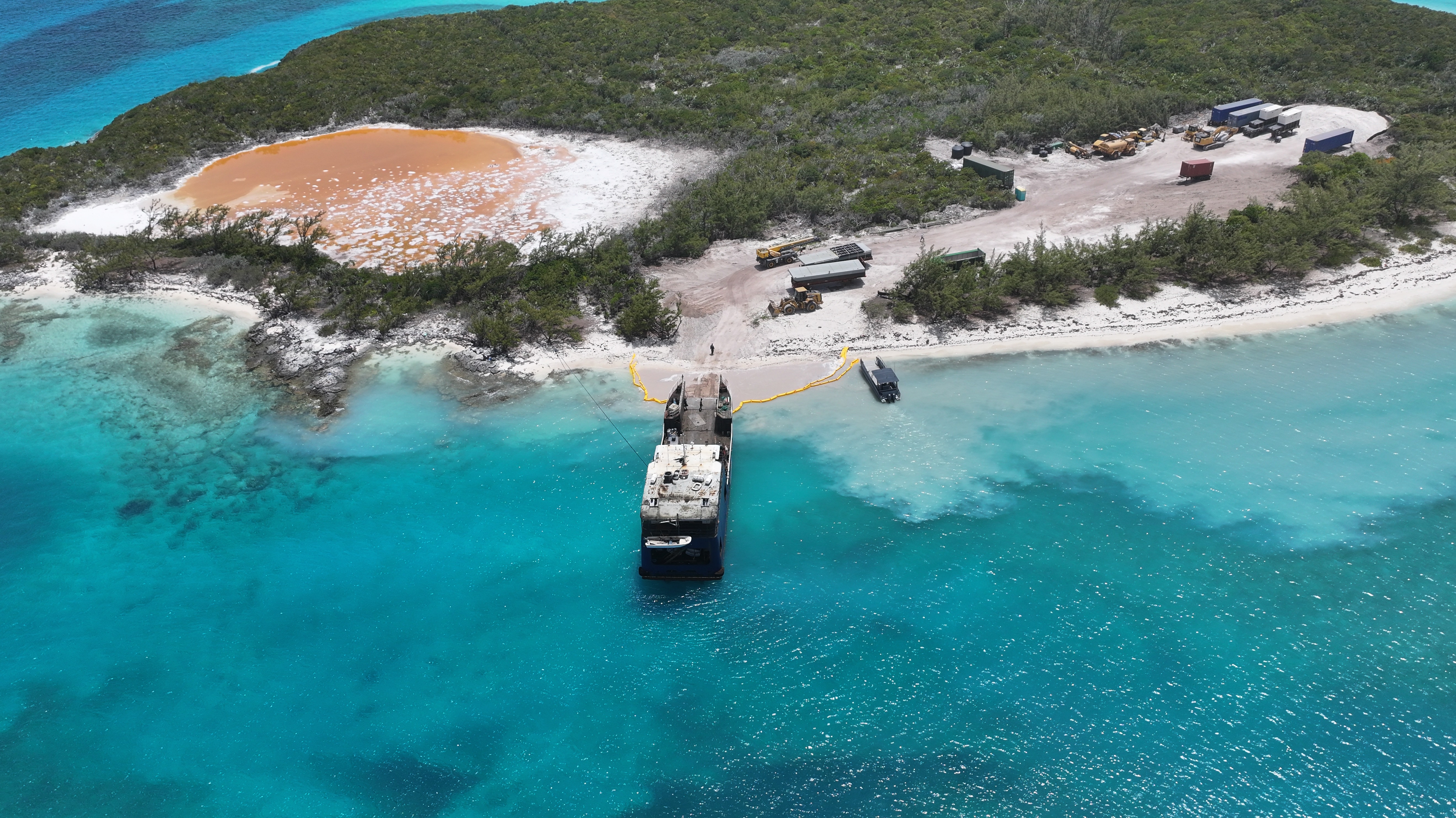 Aerial view of a ship docked at a small sandy shore on an island with dense green vegetation and turquoise water, with construction vehicles and equipment on the shore nearby.