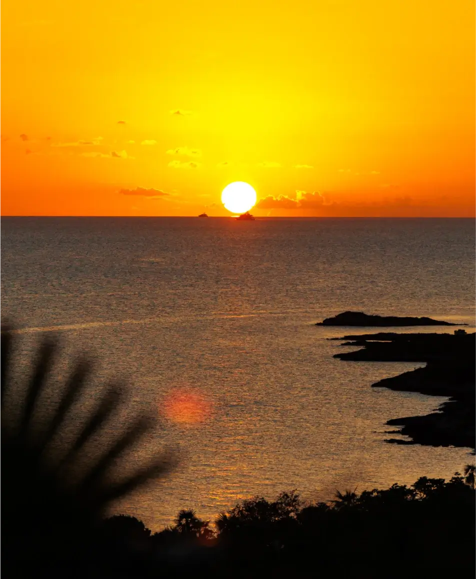 Bright orange and yellow sunset over calm ocean water with silhouetted rocky coastline and greenery in the foreground, and two boats near the horizon by the sun.