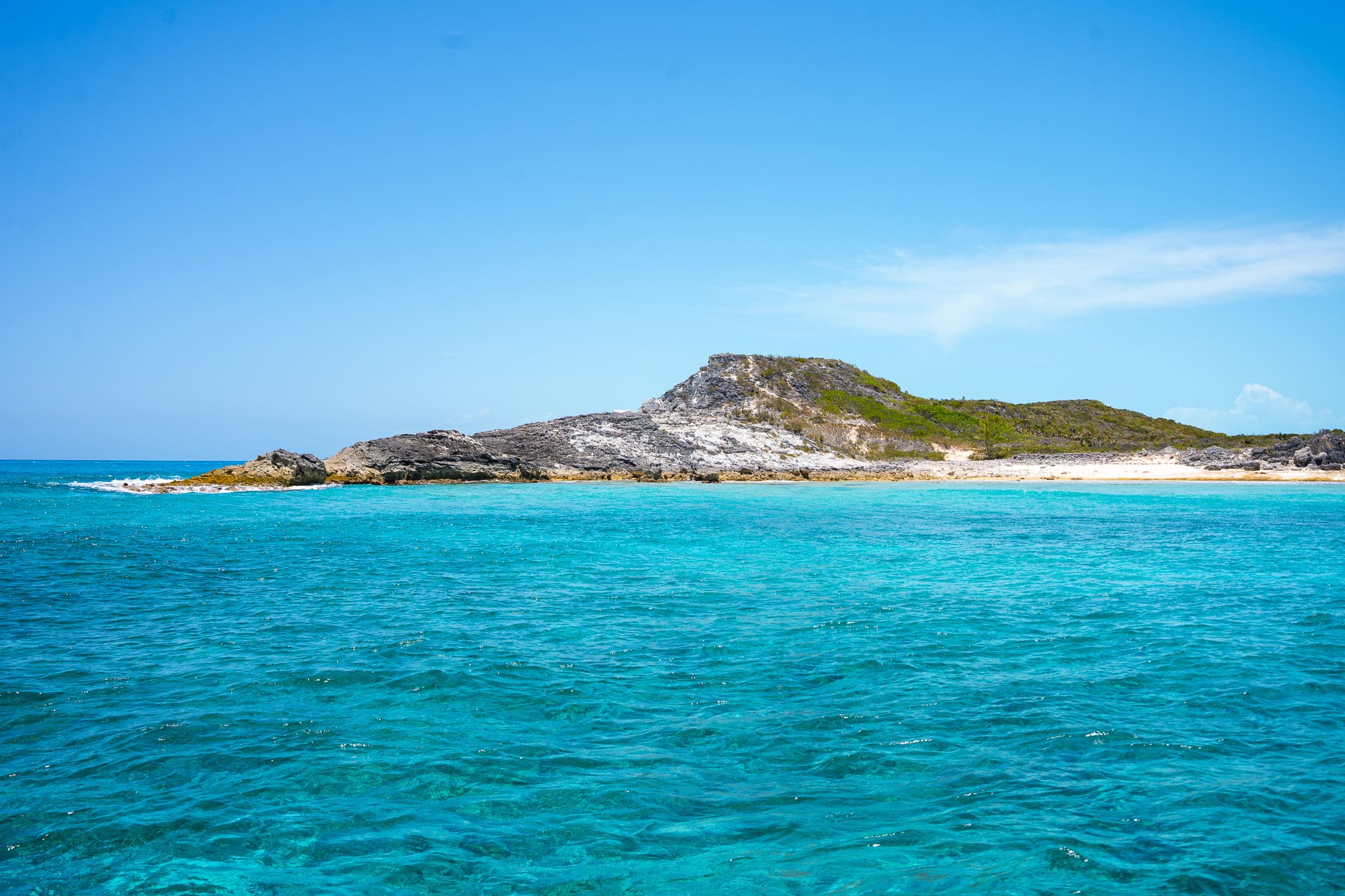 Clear turquoise ocean water in the foreground with a rocky, sparsely vegetated island under a bright blue sky in the background.