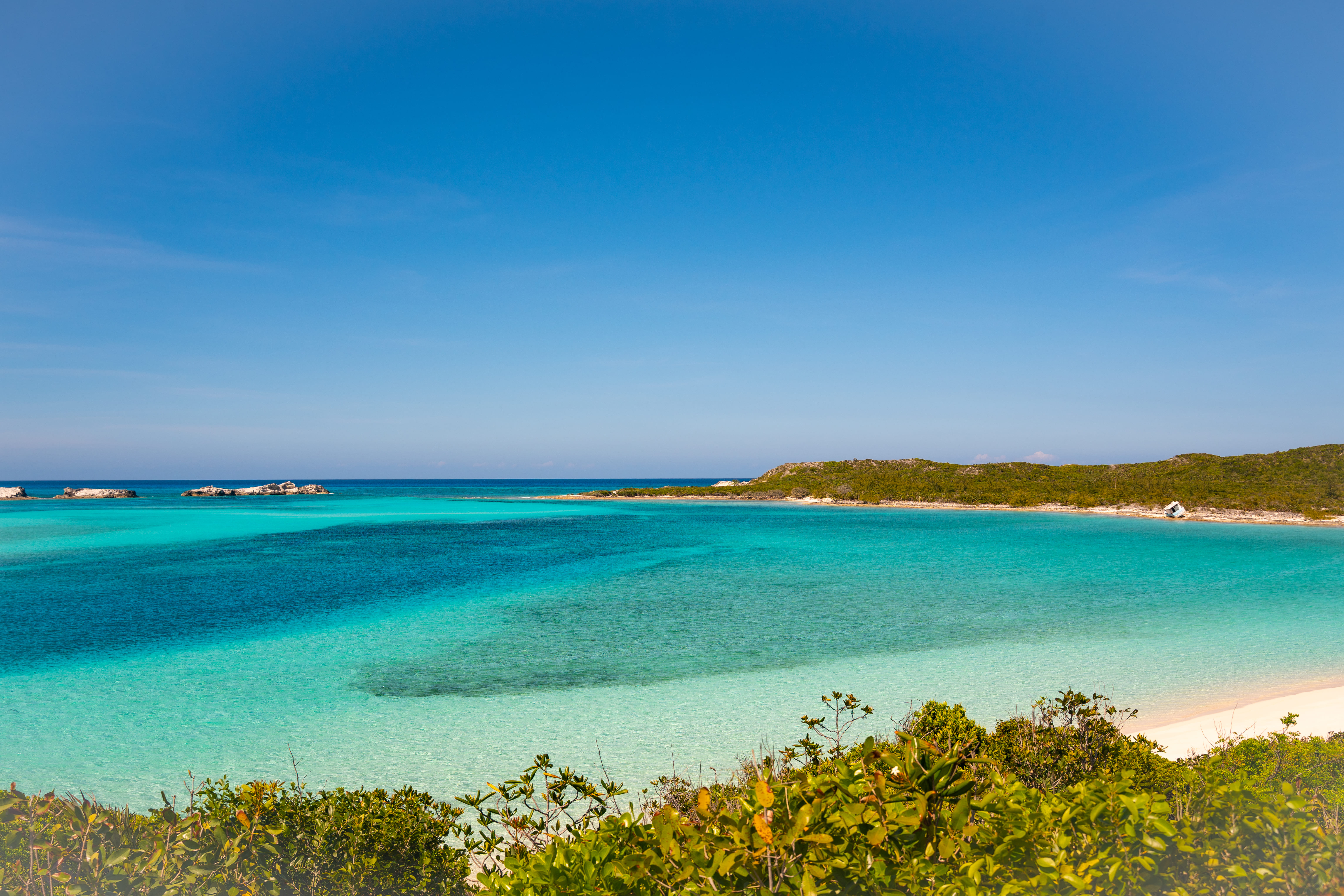 Clear turquoise water along a sandy beach with green shrubs in the foreground and a distant shoreline under a bright blue sky.