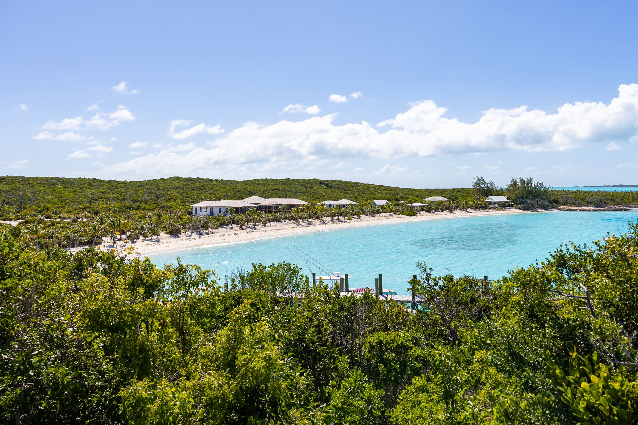 Turquoise bay with sandy beach lined by a few houses and lush green vegetation under a bright blue sky with scattered clouds.