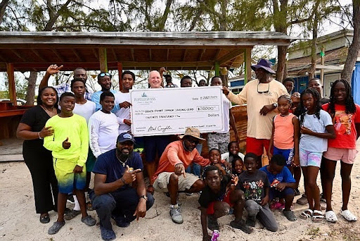 Group of diverse people of various ages outdoors, some holding a large ceremonial check for twenty thousand dollars, posing and smiling under a covered shelter with trees in the background.