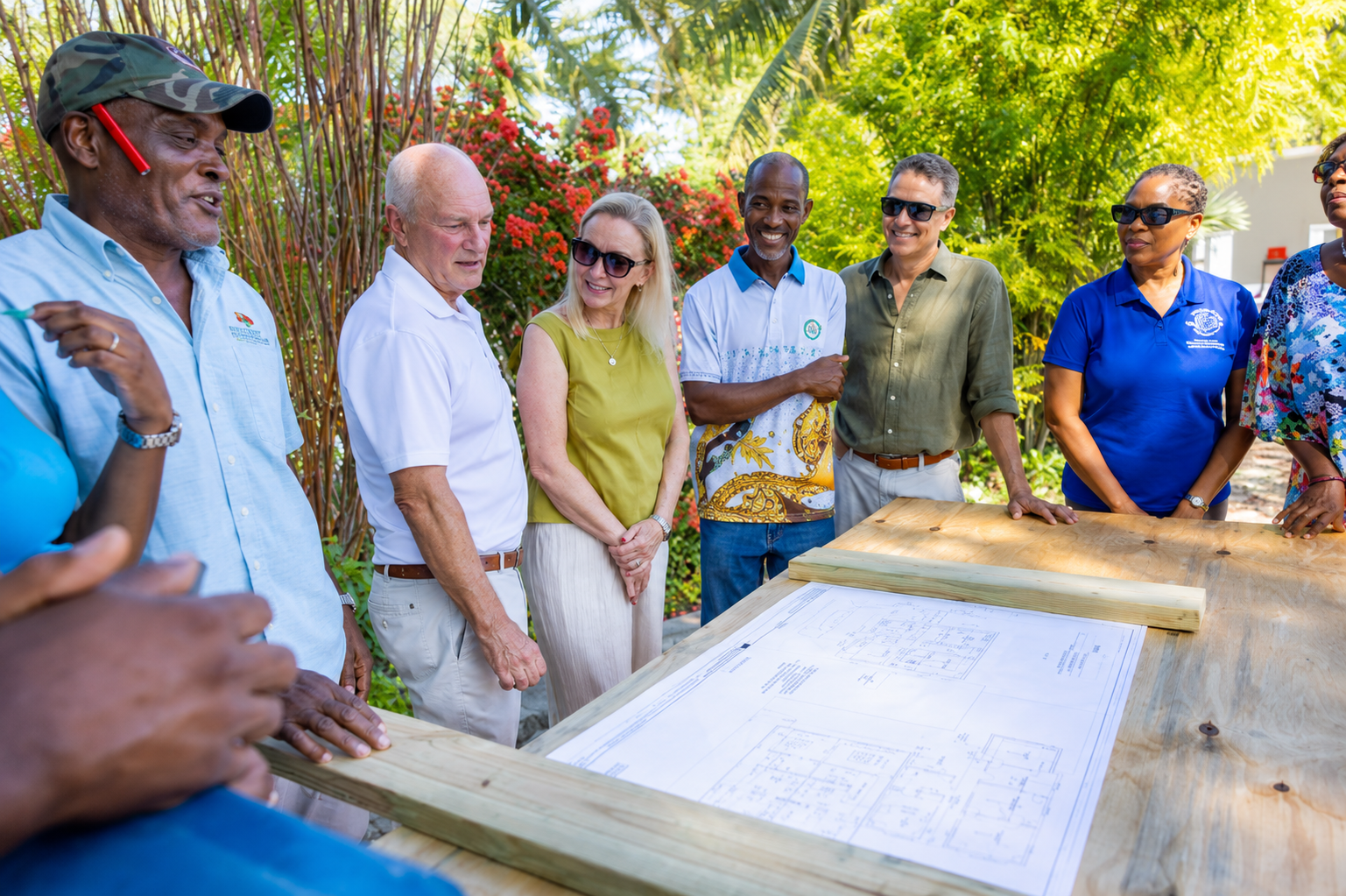 A group of six diverse adults standing outdoors around a wooden table with architectural blueprints, engaged in discussion with greenery and flowers in the background.