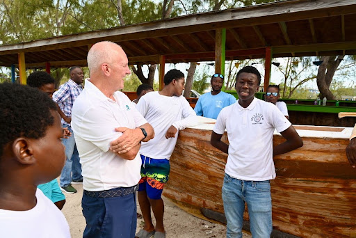 A group of people, mostly young men in casual clothing, stand outdoors near a large wooden structure under a roofed area, engaging in conversation and smiling.