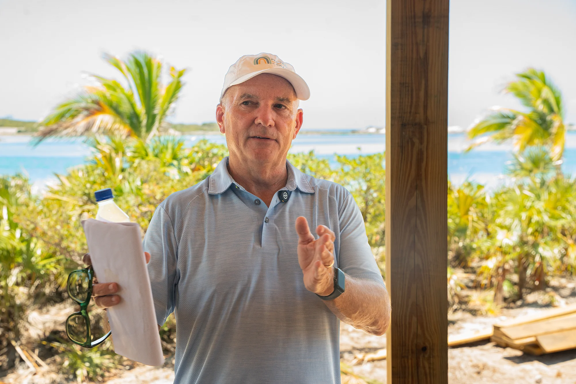 Man wearing a white cap and light blue polo shirt stands outside near tropical plants and water, holding papers with a water bottle and sunglasses in his left hand, gesturing with his right hand.