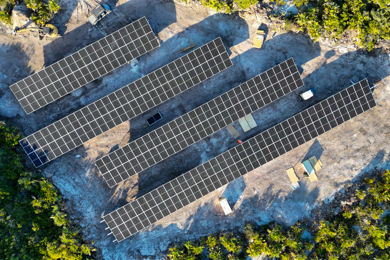 Aerial view of four long rows of solar panels installed on cleared land surrounded by green vegetation, with vehicles and an individual working nearby.
