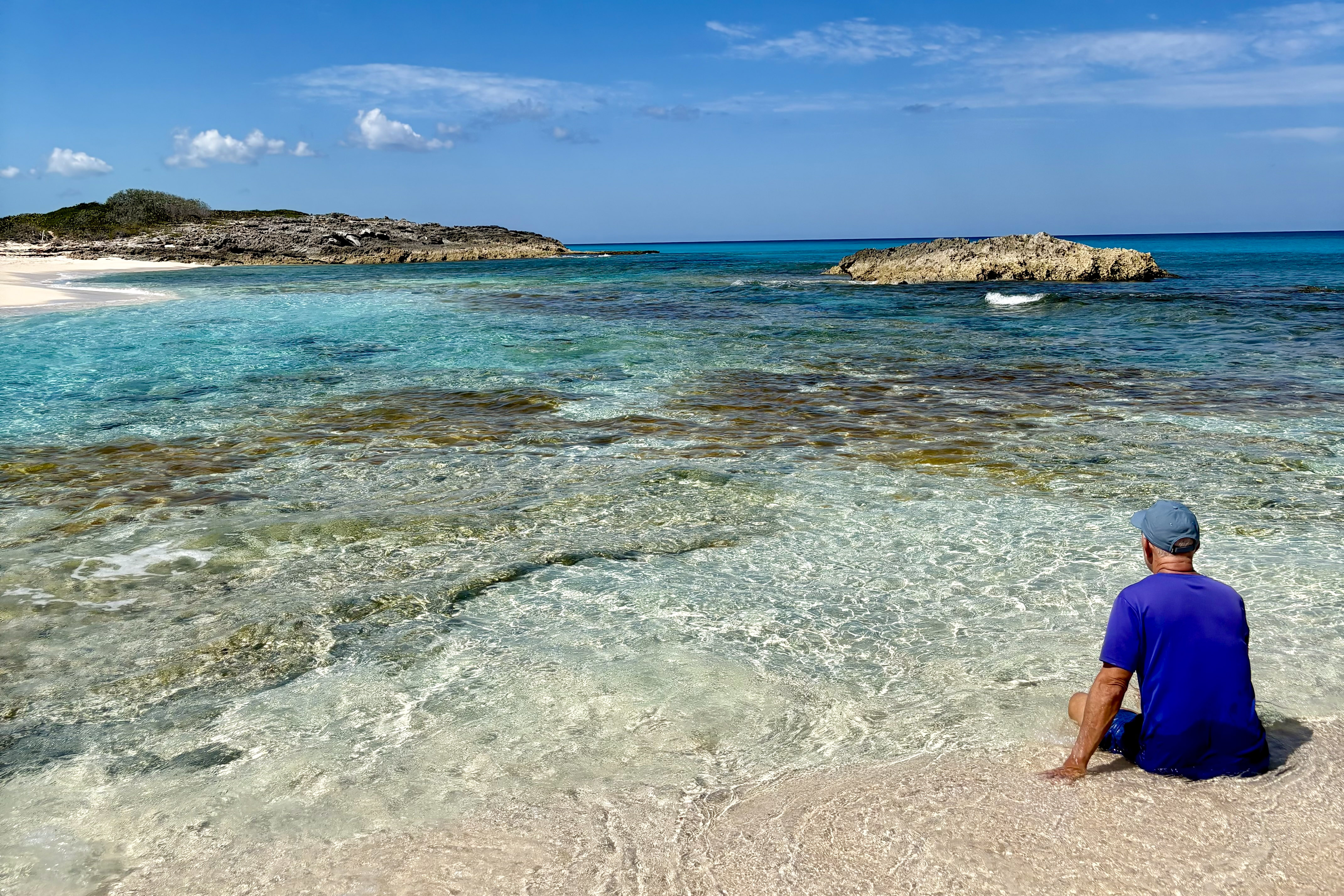 Man in blue shirt and cap sitting on sandy shore facing clear turquoise ocean with rocky islands under a partly cloudy blue sky.