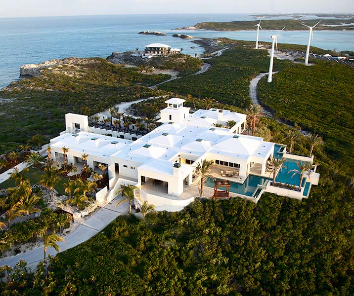 A large white modern house with multiple roofs and a swimming pool, surrounded by palm trees and greenery, set near a coastline with hills and wind turbines in the background under a clear sky.