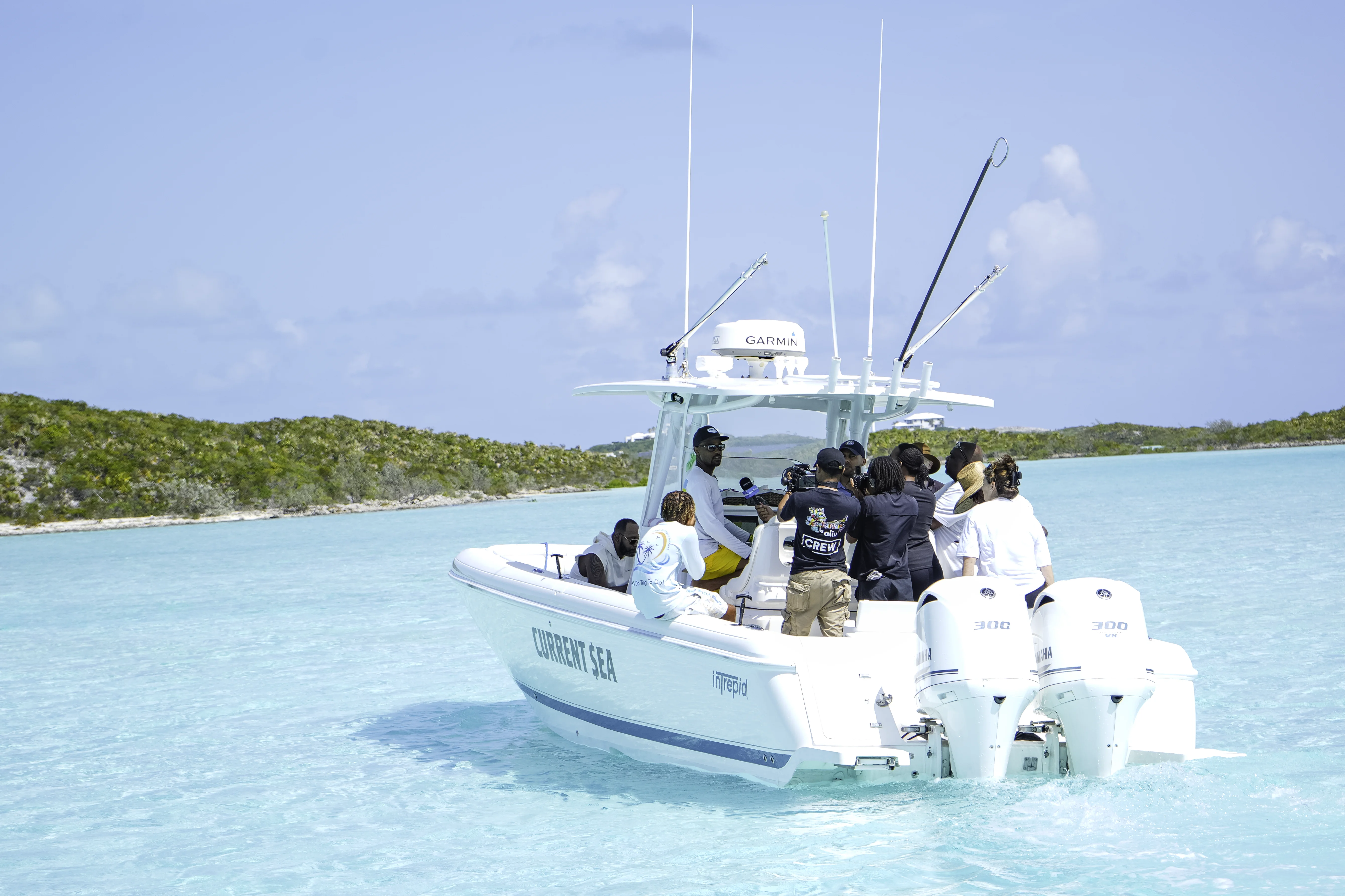 Group of people on a white boat named Current Sea in clear turquoise waters near green islands under a blue sky, with some filming and interviewing a man seated at the boat's console.