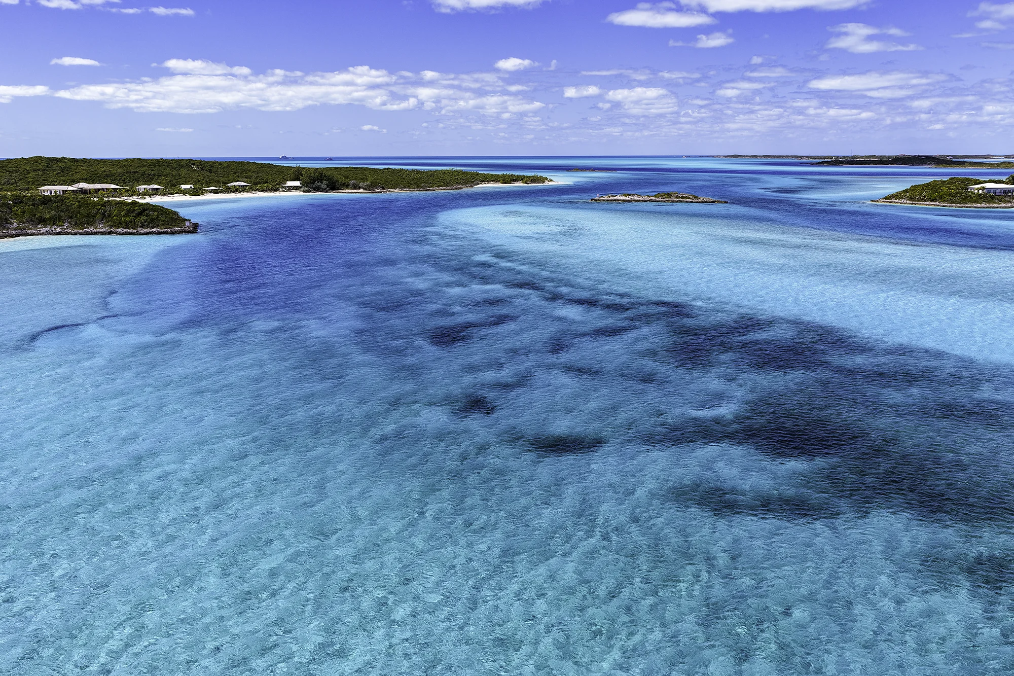 Aerial view of clear turquoise ocean water with small islands and green vegetation under a partly cloudy blue sky.
