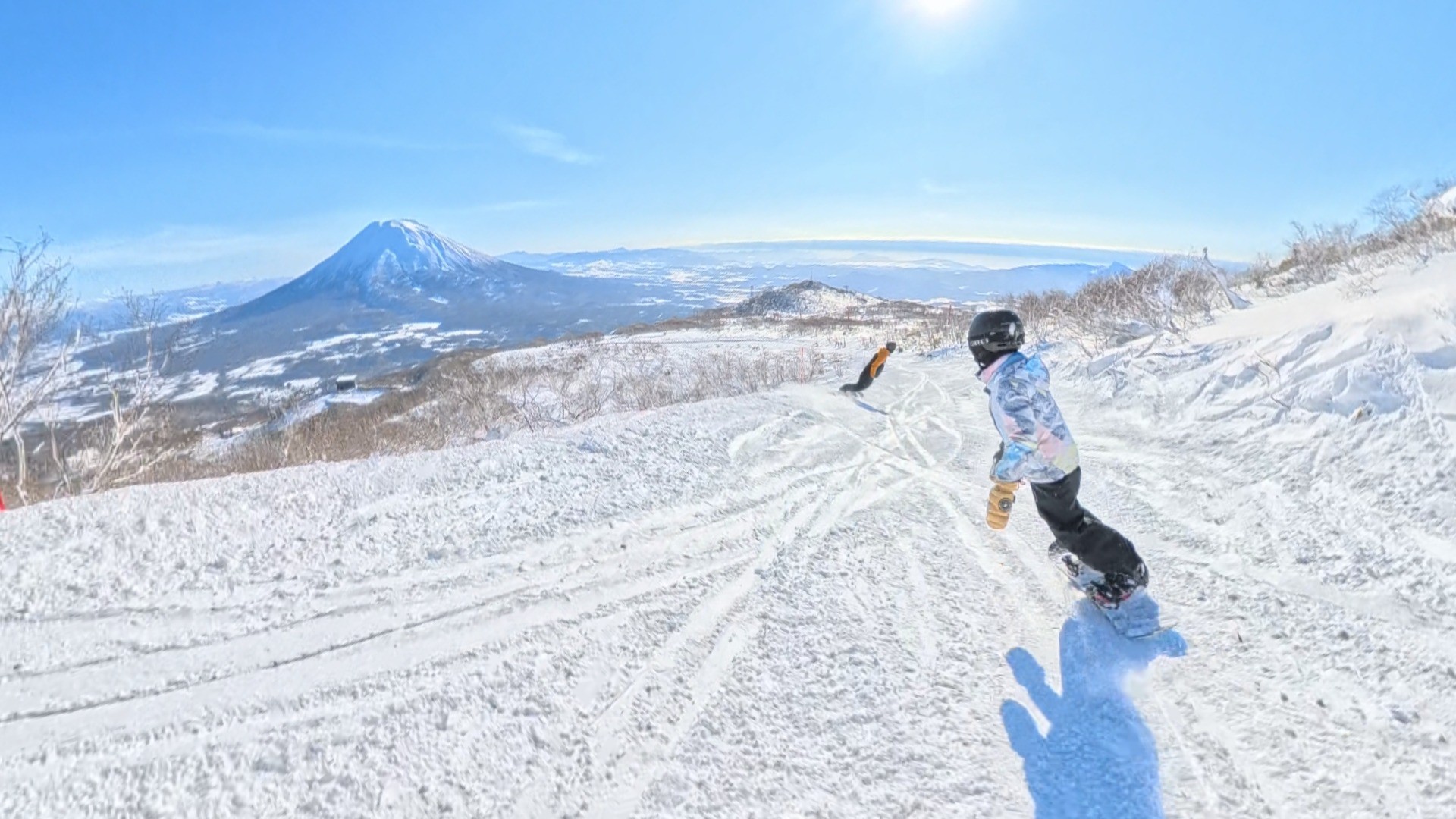 Two snowboarders descending a snow-covered mountain slope on a clear, sunny day with a large snow-capped mountain visible in the distance.