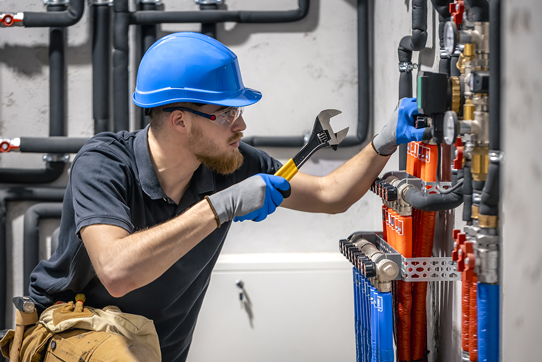 Technician wearing blue hard hat and gloves using a wrench to adjust valves on heating system pipes in a boiler room.