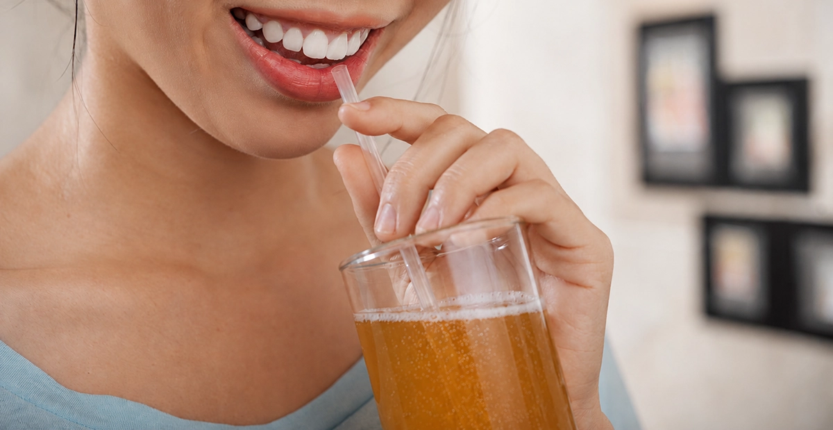 Close-up of a woman drinking kombucha through a straw, highlighting how acidic beverages can affect tooth enamel and oral health.