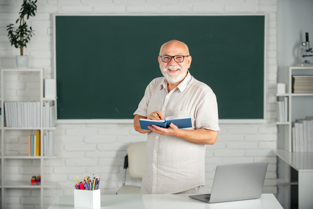 Un homme âgé debout devant un tableau noir avec un ordinateur portable.