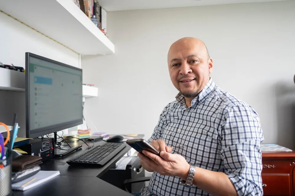 Small business owner using a smartphone beside a desktop computer in a home office