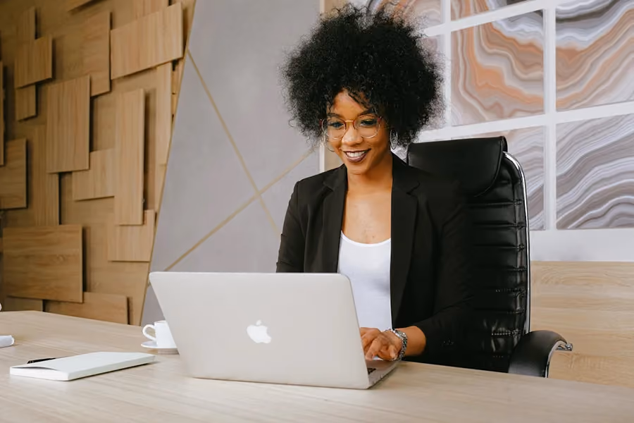 Mulher sorridente trabalhando em um notebook em um escritório moderno, com parede decorada e caderno sobre a mesa.