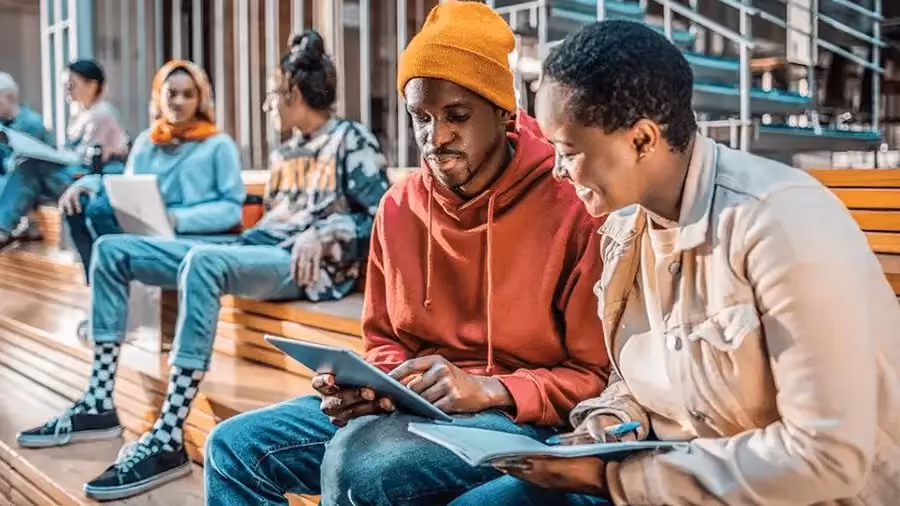 Seis pessoas jovens sentadas em uma arquibancada, em duplas, lendo no computador.