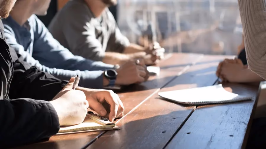 Imagem de uma mesa de reunião que mostra apenas as mãos de cinco pessoas, que estão concentradas e discutindo sobre negócios.