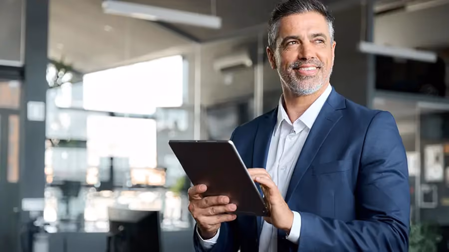 Homem com trajes formais, inspirado, sorrindo, olhando para o horizonte e segurando um tablet no ambiente de trabalho.