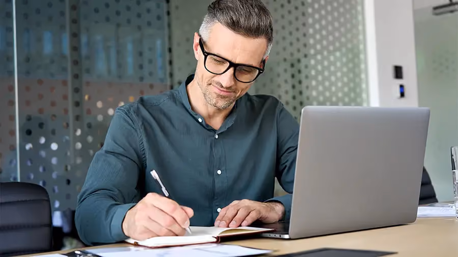 Homem sorridente fazendo anotações em uma folha de papel enquanto lê algo no seu computador.