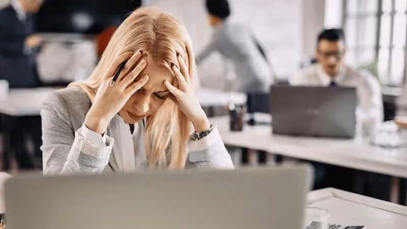 Mulher em um escritório, sentada em frente ao computador, com os braços em cima da mesa, com o rosto apoiado e irritada.