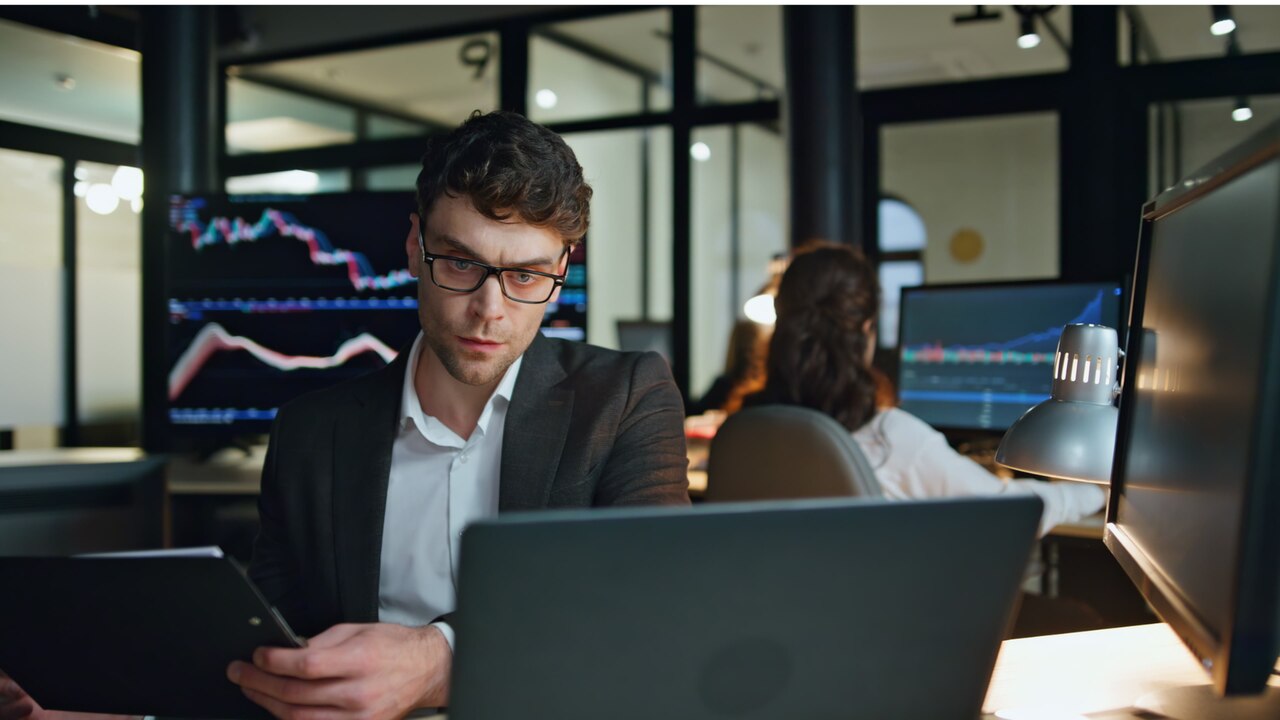 Profissional analisa dados financeiros em computador com gráficos ao fundo, representando mapeamento de risco, tomada de decisão baseada em dados e monitoramento contínuo empresarial.
