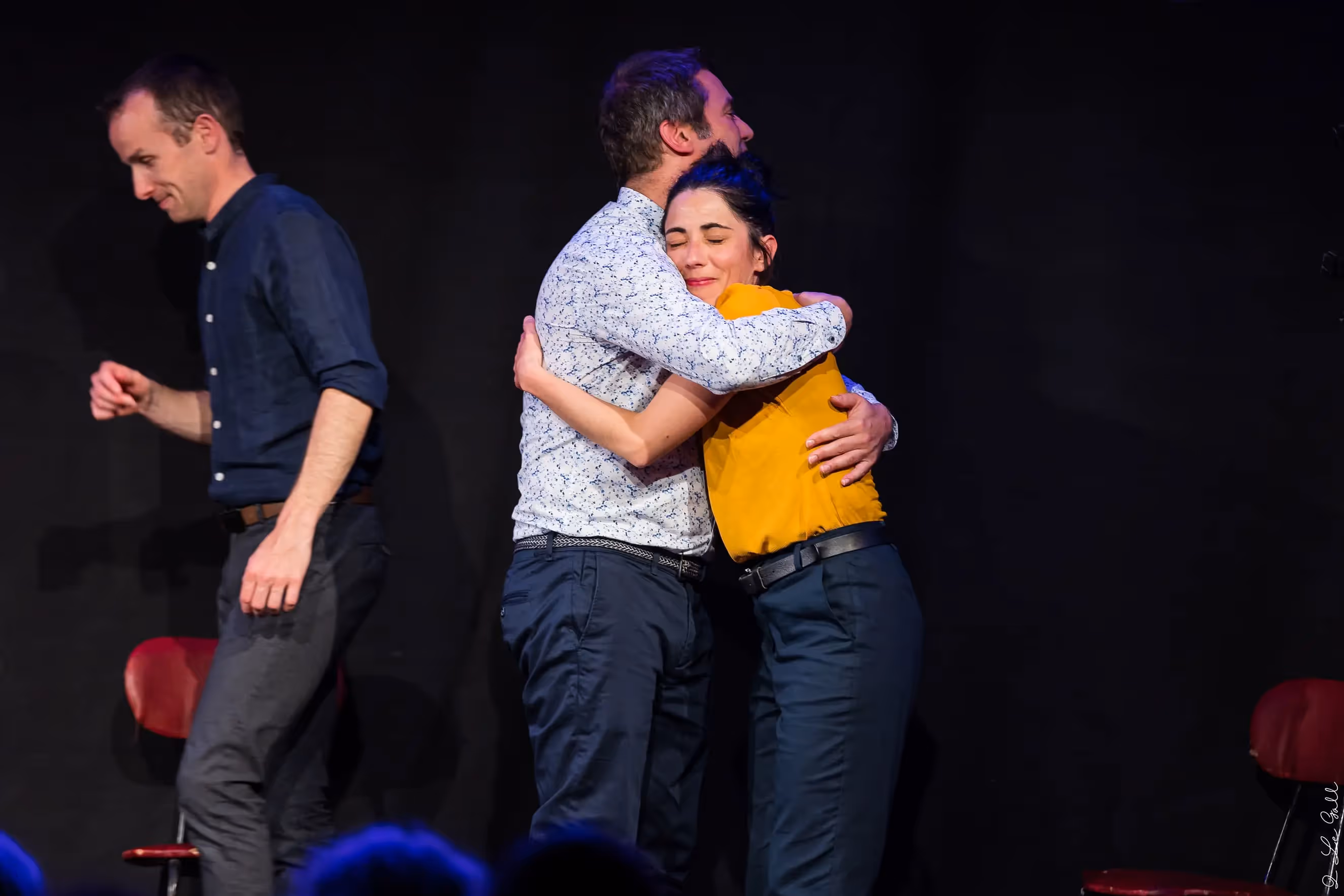 Trois acteurs en représentation théâtrale, dont deux s'enlaçant