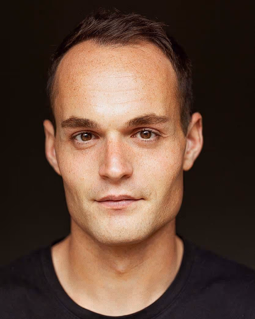 Portrait en gros plan d’un homme au visage sérieux, cheveux courts, portant un t-shirt noir, avec un fond sombre.