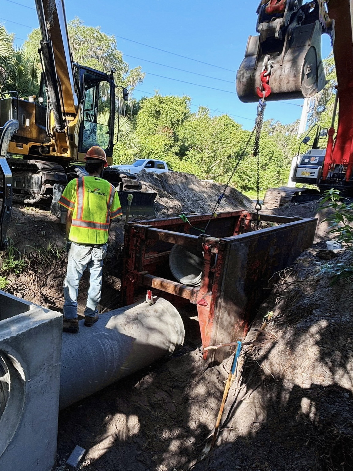 Construction worker wearing a helmet and safety vest guiding pipe installation near heavy machinery.