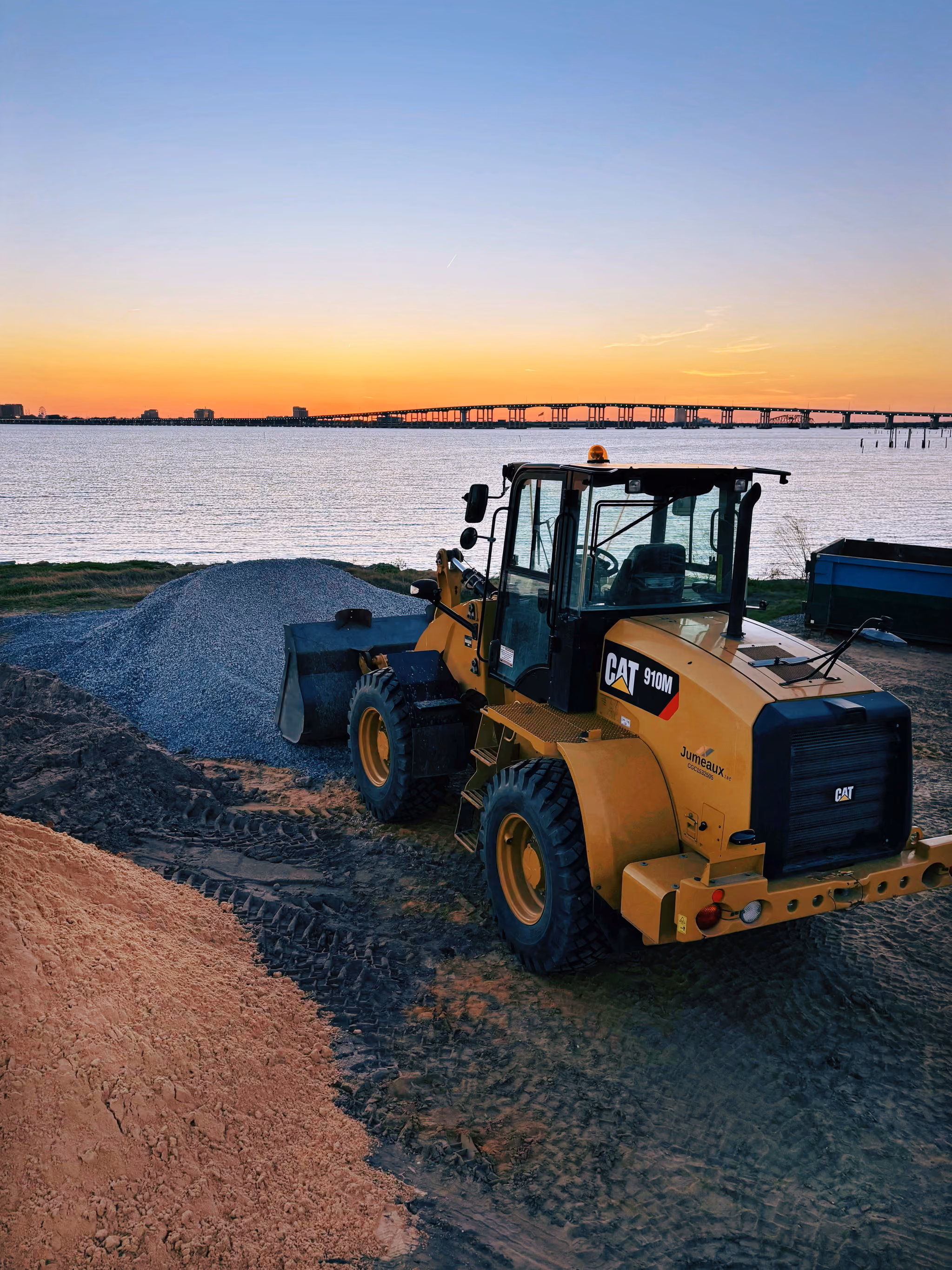 Yellow CAT 910M loader with gravel piles near waterfront at sunset with a distant bridge.