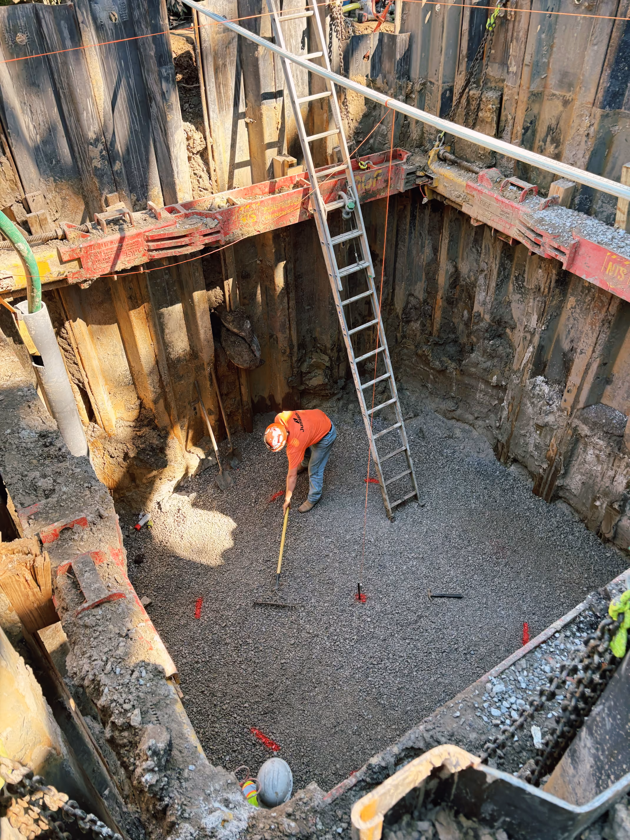 Construction worker smoothing gravel base in deep excavation supported by metal trench walls and braces.