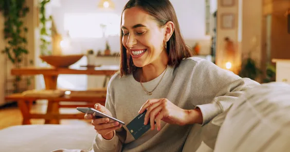 Pessoa sorrindo no sofá com o celular e um cartão de crédito