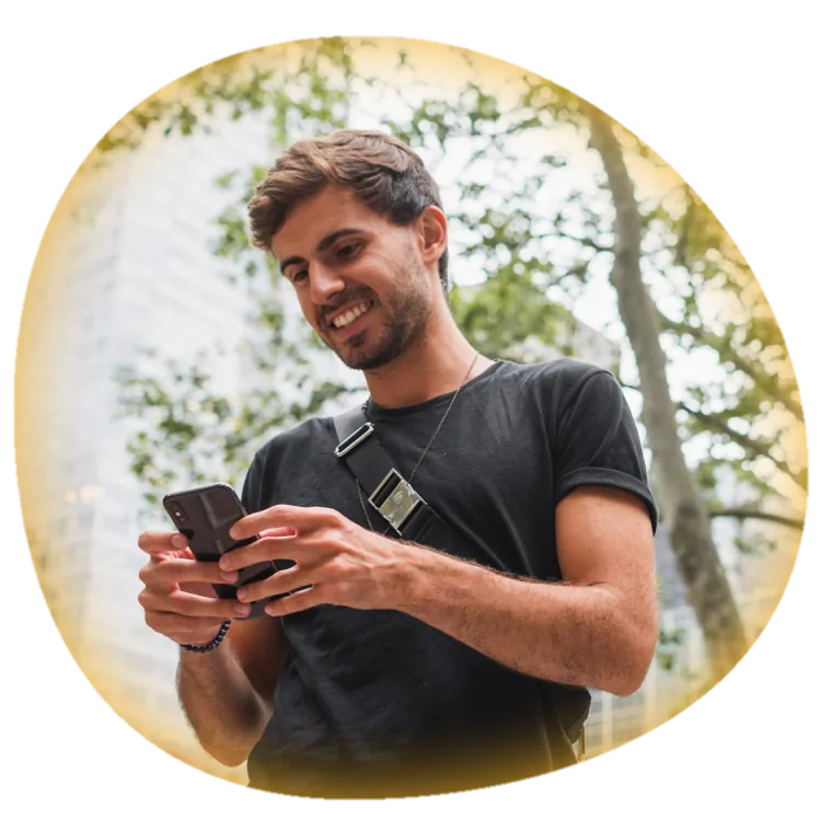 Homem jovem sorrindo enquanto usa o celular ao ar livre, com árvores e um prédio ao fundo