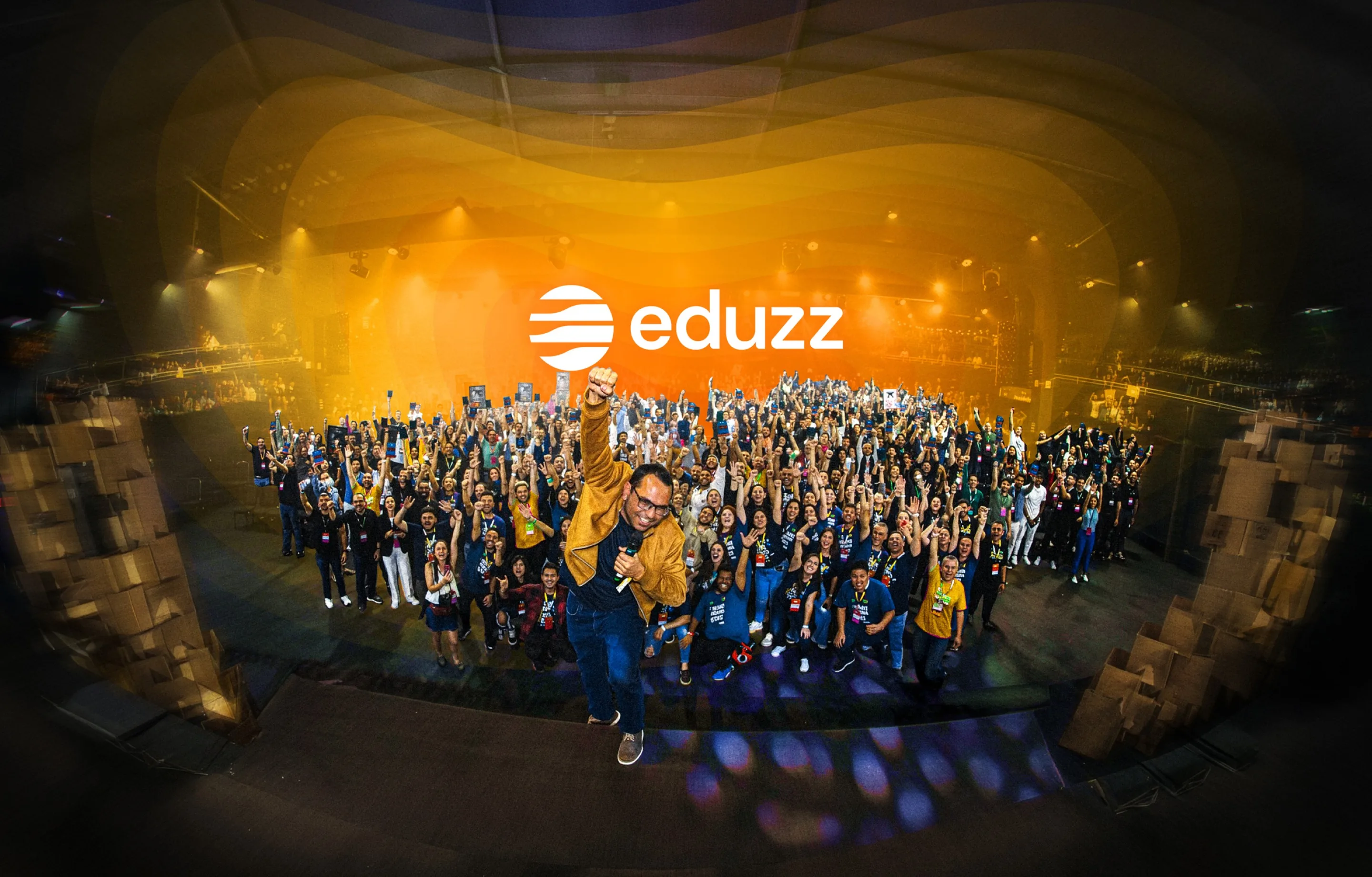 Plateia de um evento da Eduzz vista em olho de peixe, com uma pessoa de braço erguido em primeiro plano e a assinatura da marca sobre o palco